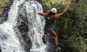 A canyoner mid-air during a jump with a waterfall in the foreground.