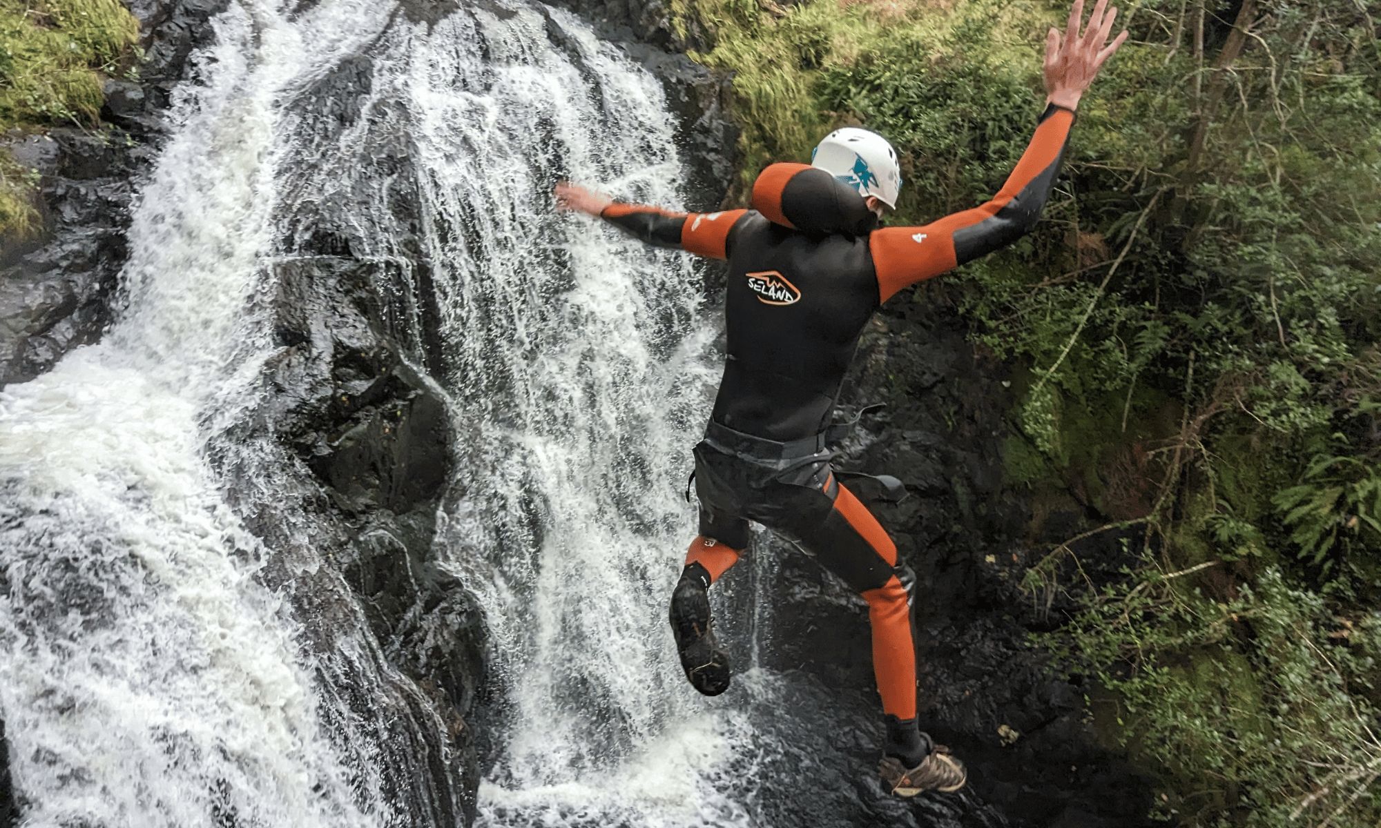 A canyoner mid-air during a jump with a waterfall in the foreground.